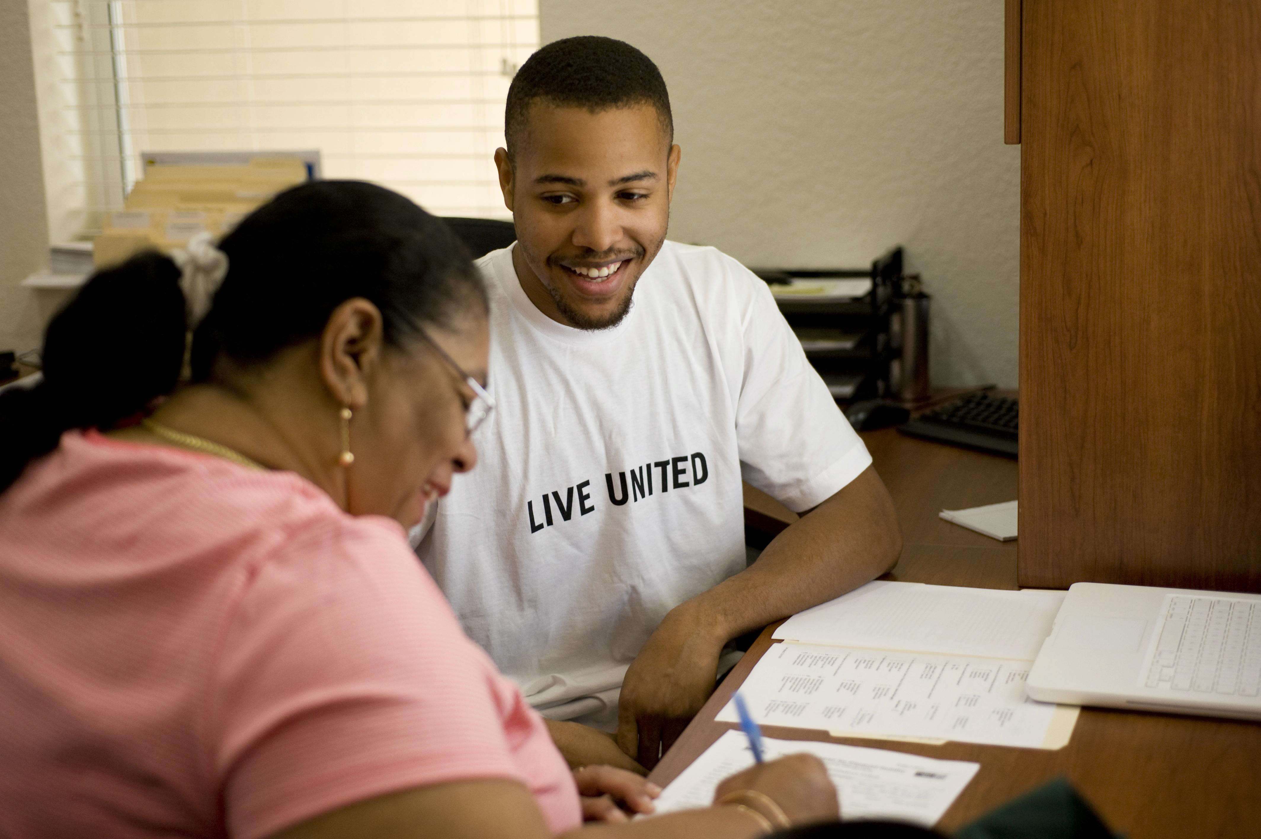 A person wearing a Live United t-shirt smiles as another fills out paperwork at a desk.