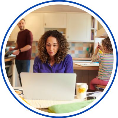 In a bright office kitchen, a woman with curly hair sits at a desk using a laptop, while a man stands in the background and a small child sits at the side.