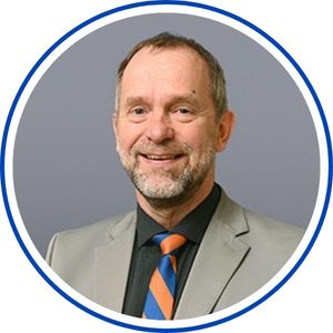 Head-and-shoulders portrait of a smiling middle-aged man in a light gray suit, dark shirt, and blue-and-orange striped tie, set against a solid gray background within a circular frame.