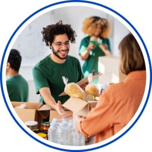 People packing and assembling food or care boxes at a table, with cardboard boxes, bread rolls, and water bottles visible; one person in a green shirt smiles while working.