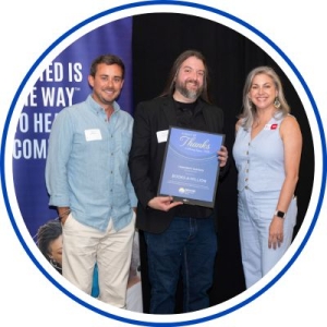 Three people stand on a stage posing for a photo; the center person holds a blue award plaque, with a blue campaign-style backdrop reading “UNITED IS THE WAY…” behind them.