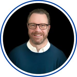 Headshot portrait of a person with short gray hair and a neatly trimmed beard, wearing dark-framed glasses and a blue sweater over a light collared shirt, against a dark background in a circular frame.