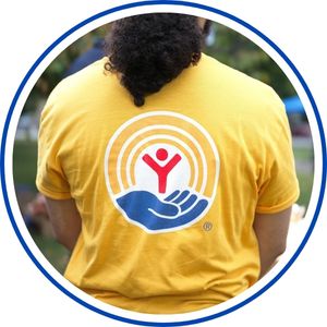 Back view of a person wearing a yellow T-shirt with a circular rainbow logo and a red hand symbol on the center, outdoors with blurred greenery in the background.