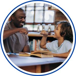 Two people sit at a table in a classroom or office, smiling and chatting while reading from open books, with blurred shelves and windows in the background.