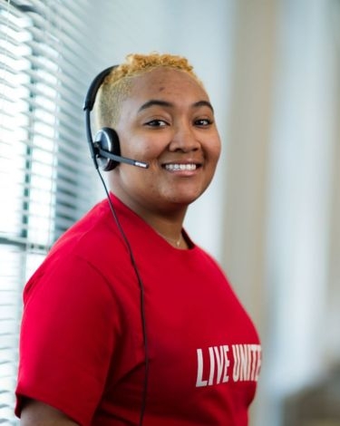 A person wearing a headset and a red t-shirt with the words "LIVE UNITED" printed on it smiles at the camera.