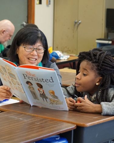 A person reads a book titled "She Persisted" to a child sitting at a desk.