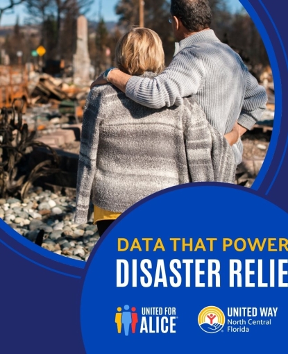 Two people stand with their backs to the camera, looking at the ruins of a house after a natural disaster. The structure is destroyed, with debris scattered around the foundation.