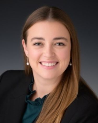A woman with long brown hair smiles widely, wearing a black blazer over a dark teal blouse and pearl earrings.