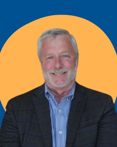 A man with gray hair and a mustache smiles at the camera, wearing a dark suit jacket over a blue checkered shirt.